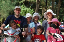 Professor James Oldroyd, his wife, and five of their children of varying ages, four girls and one young boy. They are wearing hats and riding mopeds in Thailand.