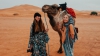 Kylie Chenn and blonde woman pose holding a rope attached to a camel. They are standing in front of a vast empty landscape of orange sand cloudy sky in the Sahara desert