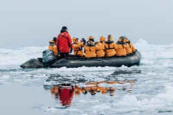 A group of Acanela travelers in orange warm coats sit in a blow up raft in the icy waters surrounding Antarctica
