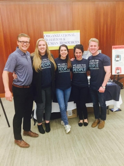 Five students stand in front of an organizational behavior and human resources booth. They are members of SHRM. There are two men and three girls.